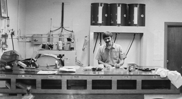 Photographer's Mate 3rd Class Terry Parkhurst at the print finishing table in the old NPTR Still Photo Lab. National Parachute Test Range, El Centro, California, circa 1974.

Photo by PHAN Pat 'Smitty' Smith.